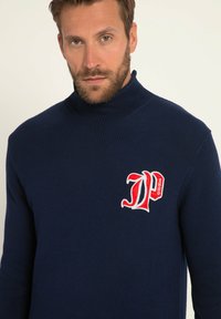 Granatowy sweter robiony na drutach z wysokim kołnierzem, z czerwonym haftowanym logo na lewej piersi. Tkanina ma teksturowane wykończenie.