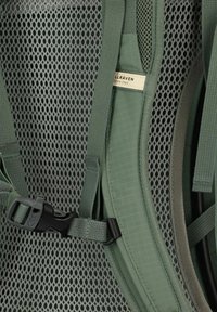 Zöld hálós és szövet hátizsák részletei, láthatók a pántok, a csat és a textúrált felület. Kiemelkedő logó címke és rácsminta is található rajta.