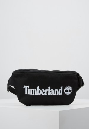 Sac banane Timberland noir avec logo blanc et symbole d’arbre, présenté sur une plateforme blanche sur un fond clair uni.