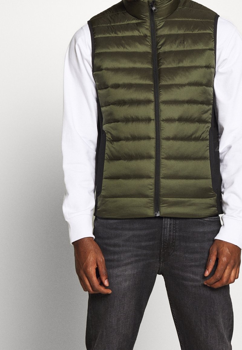 Green puffer vest with a zippered front, made of shiny fabric, featuring horizontal quilted sections and black side panels. Worn over a white sweater.