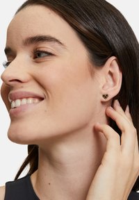 Guldtonade hjärtformade örhängen med ett bikake-mönster, uppvisade mot en modells öra, som visar en slät, polerad yta.