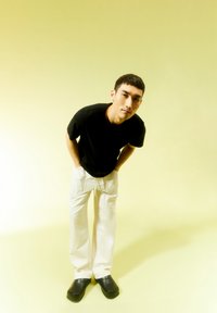 Schwarzes T-Shirt, weiße lockere Hosen und dunkle Holzklotschen. Das Modell steht leicht gebeugt mit den Händen in den Taschen vor einem hellgelben Hintergrund.