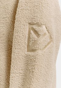 Lähikuva beige-beigestä, teksturoidusta fleecestä, jonka pinnalla on ommeltu geometrinen kuusikulmio-kuvio.