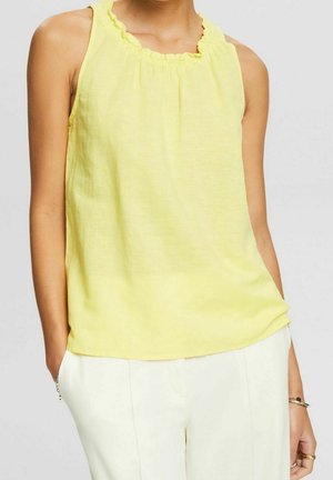 Femme portant un haut jaune sans manches avec un col volanté et un pantalon blanc, les mains dans les poches devant un fond uni.