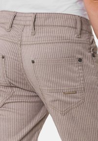 Gestreifte braune Hose mit einem taillierten Design, ausgestattet mit Gesäßtaschen mit Nieten und einem kleinen Markenetikett nahe dem Bund.