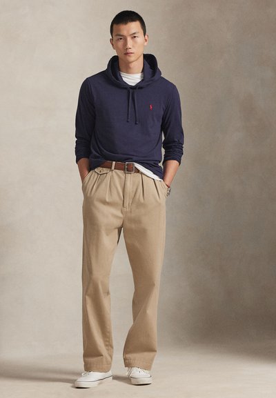 Noormees seisab käed taskutes, seljas tumesinine kapuutsiga pusa, valge särk, beežid püksid, pruun vöö ja valged vabaajajalatsid ühevärvilise tausta ees.