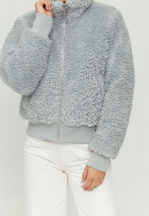 Lichtgrijs, zachtachtig jack met ritssluiting, geribde manchetten en zoom, gedragen met witte broek, hand bij de kraag om de rits aan te passen.