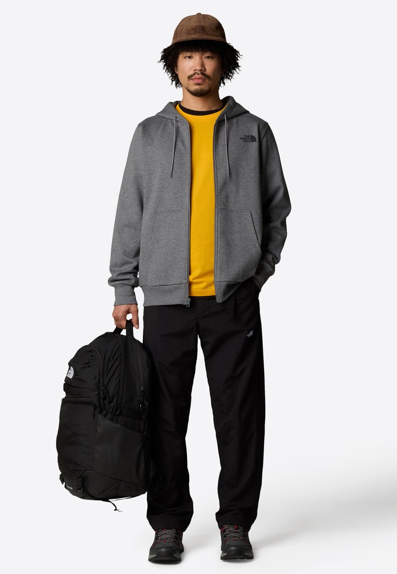 Sudadera gris con cremallera, camiseta amarilla, pantalones negros y mochila negra. Zapatillas deportivas con detalles en gris y rojo completan el atuendo.