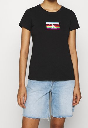 Person iført sort t-shirt med korte ærmer med Calvin Klein-logo i regnbuens farver og lyseblå denimshorts med frynset kant.