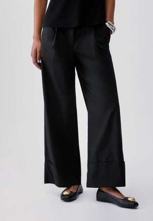 Persona che indossa pantaloni neri a gamba larga, ballerine nere con dettagli dorati, una maglia nera e braccialetti d'argento a un polso.