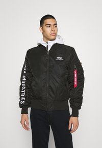 Giacca bomber nera con chiusura a zip, polsini e vita a coste. Presenta il marchio bianco sulle maniche e un'etichetta rossa sulla tasca.