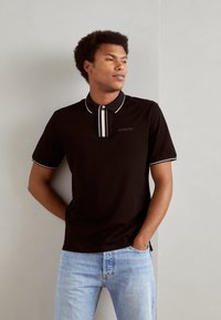 Camiseta polo negra con cuello, con acentos en blanco y una fila de botones. Lleva puesta con jeans de mezclilla azul claro. Textura de tela suave.