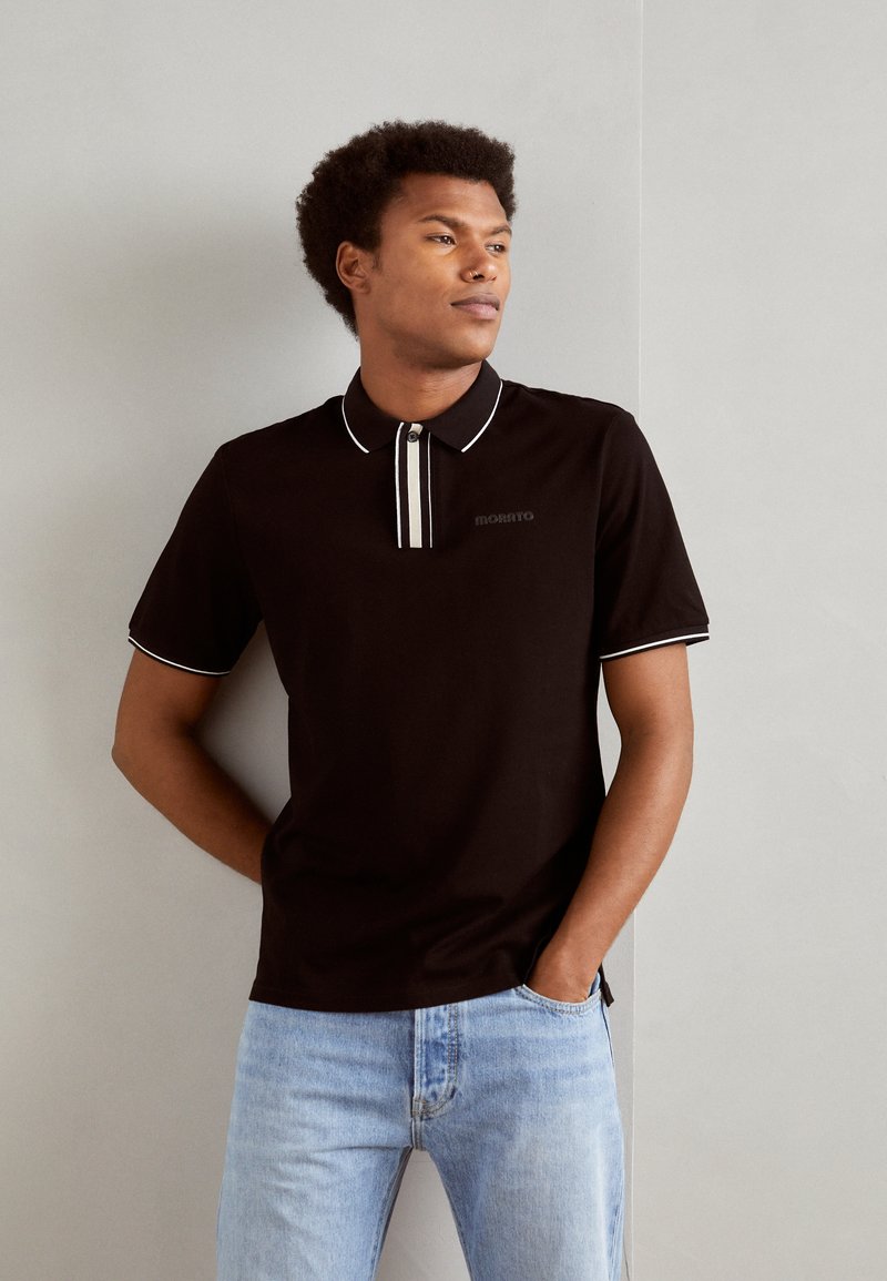 Camiseta polo negra con cuello, con acentos en blanco y una fila de botones. Lleva puesta con jeans de mezclilla azul claro. Textura de tela suave.
