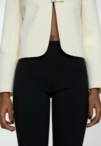 Blazer cropped crème avec un design structuré et un fermoir central, associé à un pantalon noir moulant. Texture lisse, coupe ajustée.