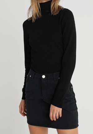 Femme portant un col roulé noir à manches longues rentré dans une jupe en jean noire avec des poches avant et une fermeture par bouton argenté.