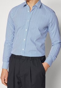Camisa de botones en azul y blanco, con un patrón geométrico. Mangas largas, cuello puntiagudo y una textura de tela suave.