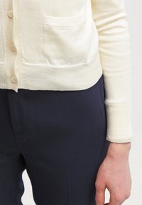 Cárdigan crema con botones, que presenta un dobladillo redondeado y un bolsillo. Combinado con pantalones azul marino, el tejido parece suave y liso.