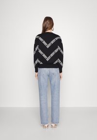 Schwarzer Pullover mit wiederholtem weißen "DKNY"-Logo im diagonalen Muster, kombiniert mit hellblauen geraden Jeans mit fransigen Säumen.