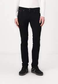 Jean skinny noir en denim, avec une poche avant ornée d'un accent rouge et blanc. La coupe est ajustée, avec une texture lisse.