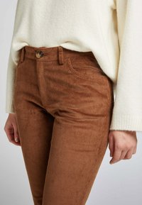 Pantalons en corduroy marron au design ajusté, avec poches avant et fermeture à bouton. Associés à un pull en tricot couleur crème.