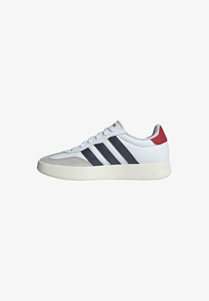 Zapatillas blancas con rayas gris y marino, con lengüeta acolchada y acento en el talón rojo. La parte superior es de cuero, con una puntera texturizada y suela de goma.