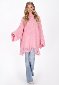 Liela izmēra rozā adīta pončo ar augstu apkakli, plašām piedurknēm un dekoratīvām maliņām. Apvienots ar platām zilo džinsu biksēm un gaišiem apaviem.