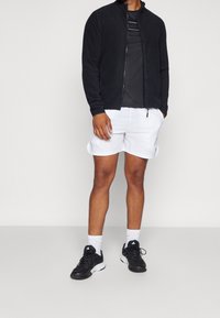 Hombre vestido con una chaqueta negra con cremallera, camiseta negra, pantalones cortos blancos, calcetines blancos y zapatillas Adidas negras, de pie con las manos en los bolsillos.