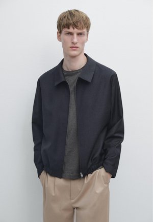 Jonge man met lichtbruin haar draagt een donkergrijs jack met rits over een antracietkleurig shirt en beige broek, staand tegen een effen witte muur.