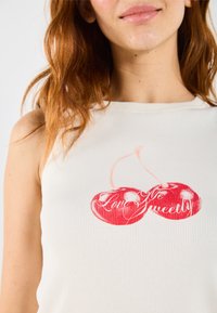 Mouwloos ribgebreid topje in lichtbeige met een rode kersafbeelding en de tekst "Love Sweetly". Soepele structuur, aansluitend ontwerp.