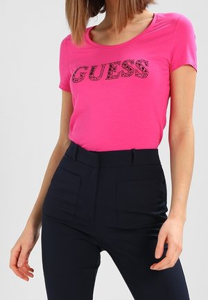 Vrouw in een felroze T-shirt met korte mouwen met het "GUESS" logo en hoge donkerblauwe broek met voorkruiken.