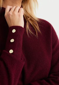 Burgundské pletené svetr s vysokým límcem a třemi knoflíky ve zlatém tónu na manžetě. Měkká textura s volným střihem.