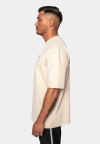 Beige bomulds t-shirt med korte ærmer, rund hals, løs pasform, vist fra siden. Har et enkelt design og synlig tatovering på armen.