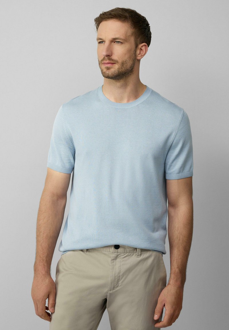 Lichtblauwe gebreide trui met korte mouwen en een ronde halslijn, met geribde manchetten, gecombineerd met beige broek, tegen een neutrale achtergrond.