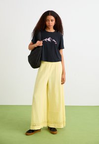 Zwarte T-shirt met korte mouwen en een roze bergafbeelding, gecombineerd met losse gele wijde broek met kantdetails, en een zwarte gewatteerde tas.