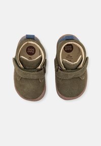 Sapatos de camurça verde com sola castanha, apresentando uma tira de velcro e um destaque azul na parte de trás, adequados para crianças.