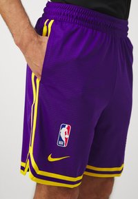 Vijolične košarkaške hlače z rumenimi poudarki, ki vključujejo logo NBA in Nike žig. Narejene iz teksturirane tkanine, zasnovane za udobje.