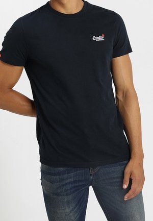 Homme portant un t-shirt noir ajusté Superdry et un jean bleu, debout avec un bras plié derrière le dos.