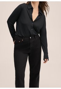 Camisa negra de manga larga con botones y cuello, combinada con unos vaqueros negros de cintura alta que tienen acentos de cobre en los bolsillos. Tejido suave.