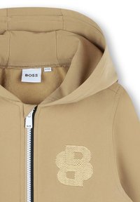 Giacca cappuccio beige con zip frontale, dettagli a costine e un logo ricamato prominente sul petto. Tessuto morbido.