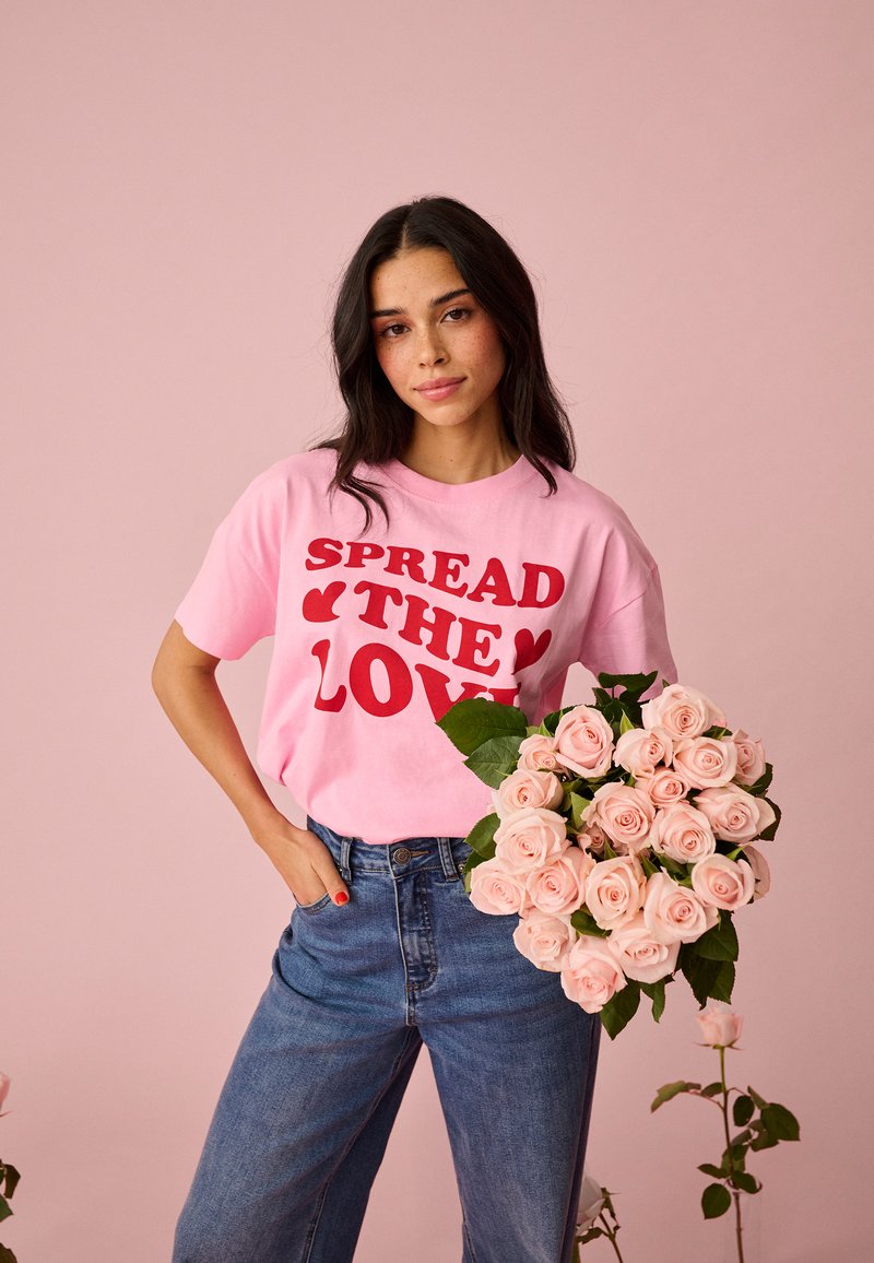 Mladá žena s dlhými tmavými vlasmi drží kyticu bledoružových ruží, má na sebe ružové tričko s nápisom "SPREAD THE LOVE" a modré džínsy.