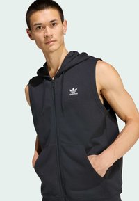 Crna kapuljača bez rukava s patentnim zatvaračem, izrađena od mekanog materijala, s prednjim džepom i bijelim Adidas logom na lijevoj strani prsa.