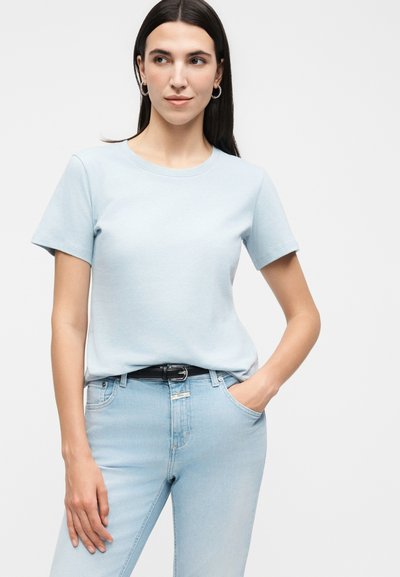 Γυναίκα με μακριά σκουρόχρωμα μαλλιά, που φοράει γαλάζιο κοντομάνικο μπλουζάκι, γαλάζια τζιν, μαύρη ζώνη και κρίκους, ποζάροντας με το ένα χέρι στην τσέπη.