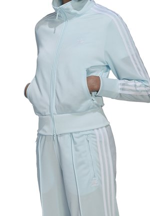 Osoba ubrana w jasnoniebieski dres Adidas z białymi paskami, z rękami w zamykanych kieszeniach, stojąca na białym tle.