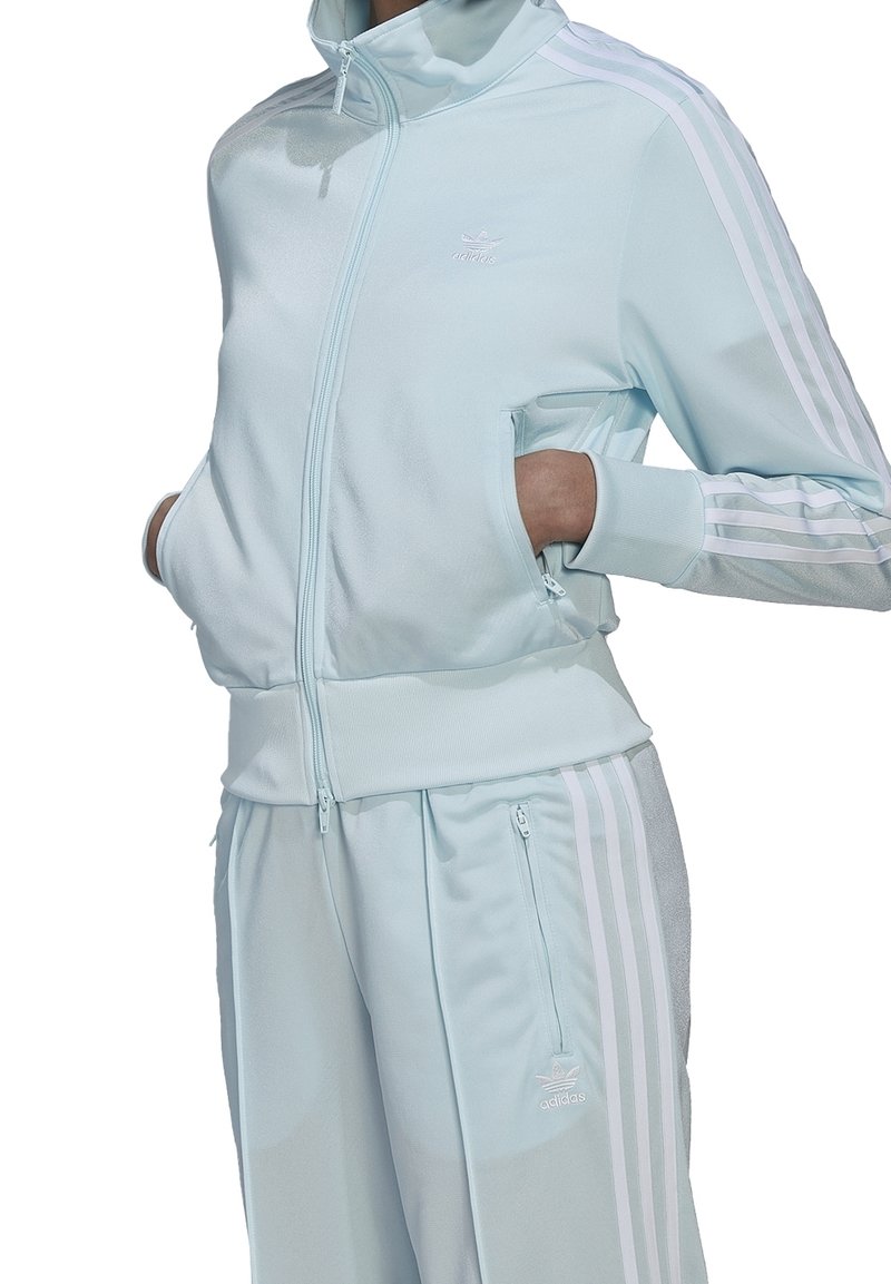 Persona con un chándal ligero de Adidas en color azul claro con rayas blancas, con las manos en los bolsillos con cremallera, de pie frente a un fondo blanco.