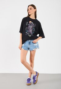 Tricou negru cu mânecă scurtă, cu un grafic colorat de dragon, asortat cu pantaloni scurți din denim uzat și teniși purpurii cu dungi albe.