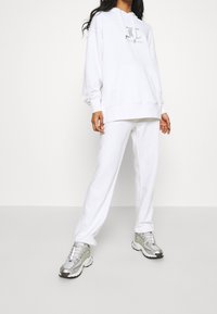 Witte hoodie met logo op de borst, gecombineerd met bijpassende witte sweatpants. Model draagt zilveren sneakers met opvallende patronen en texturen.