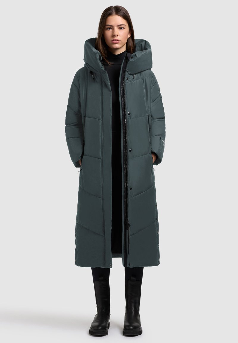 Duga pufasta jakna tamnosive boje, s visokim ovratnikom, zatvaranjem na zip i kopčom, lijepim prošivom i bočnim džepovima. Glatka, vodootporna tkanina.