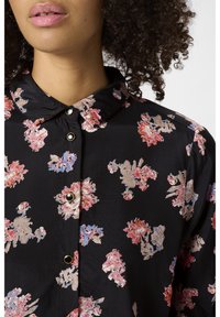 Kwiatowa bluzka z guzikiem w czarnym materiale, z różowymi, niebieskimi i beżowymi wzorami kwiatowymi, z wyraźnym kołnierzykiem i czarnymi guzikami.