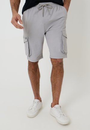 Mand iført lysegrå cargoshorts med snøre i taljen og hvide sneakers, stående mod en ensfarvet baggrund.