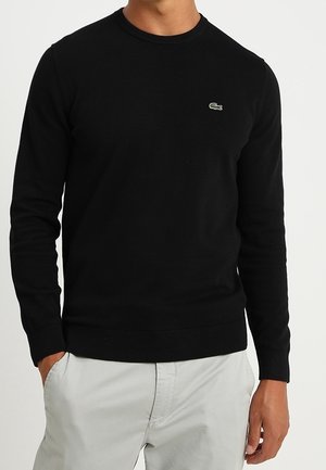 Ein Mann trägt einen schwarzen Langarmpullover mit kleinem grünem Krokodil-Logo auf der Brust, kombiniert mit hellgrauen Hosen, Hände in den Taschen.